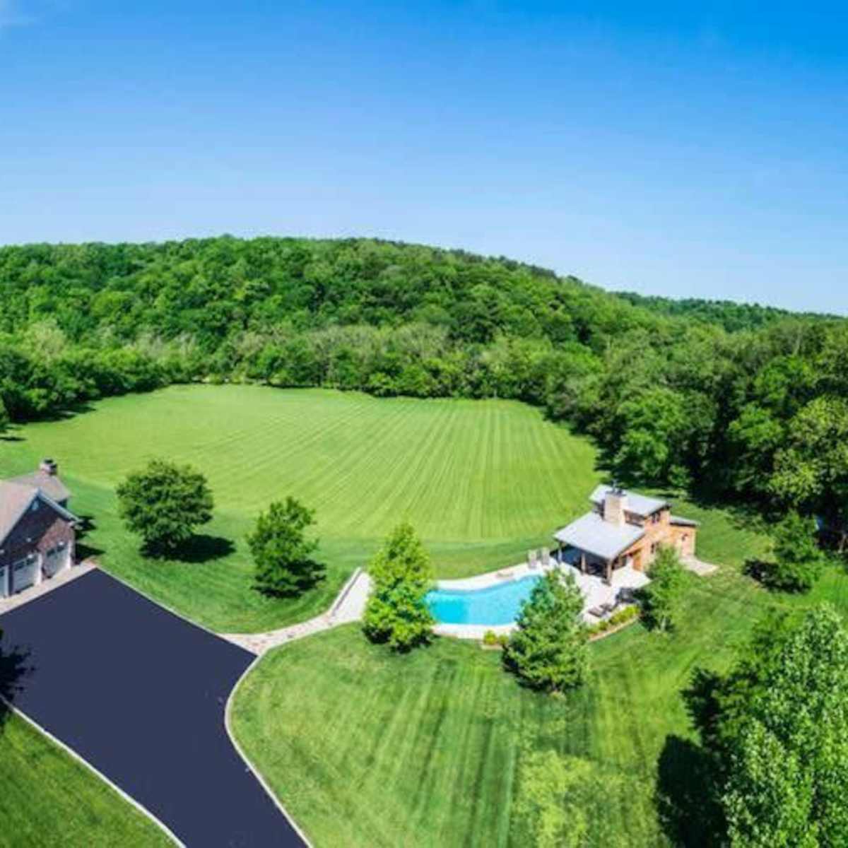 A large house with a pool surrounded by a manicured lawn and wooded hills in the background.