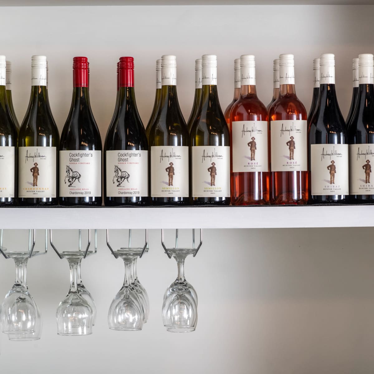 A shelf displays a variety of wine bottles in different colors and labels, alongside hanging wine glasses.