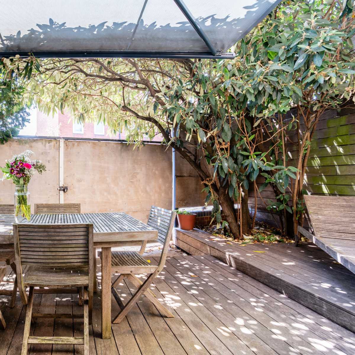 A wooden patio table with several chairs is set under a canopy, surrounded by greenery and potted plants.