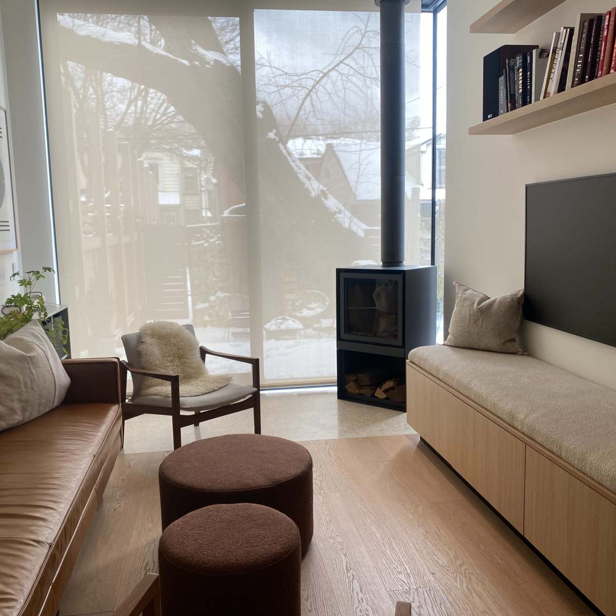 A modern living room with a leather sofa, two round stools, a wooden bench with a cushioned top, and a large window covered by sheer curtains that reveal a snowy outdoor scene.
