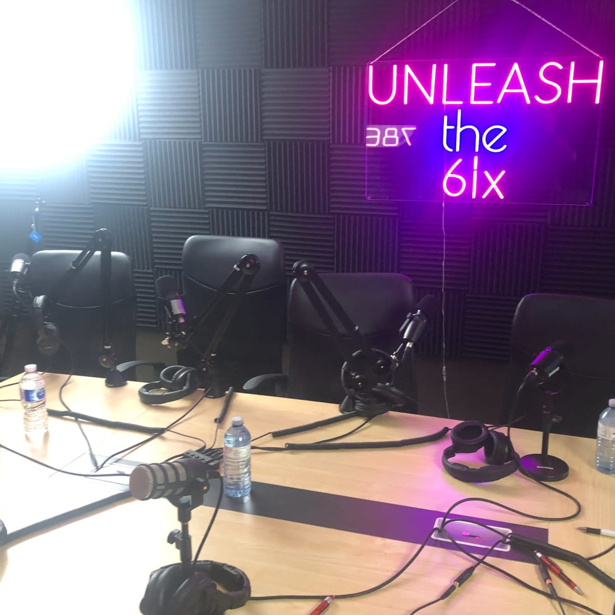 A microphone setup is arranged around a table in a podcast recording studio with a neon sign that reads "UNLEASH the 6ix."