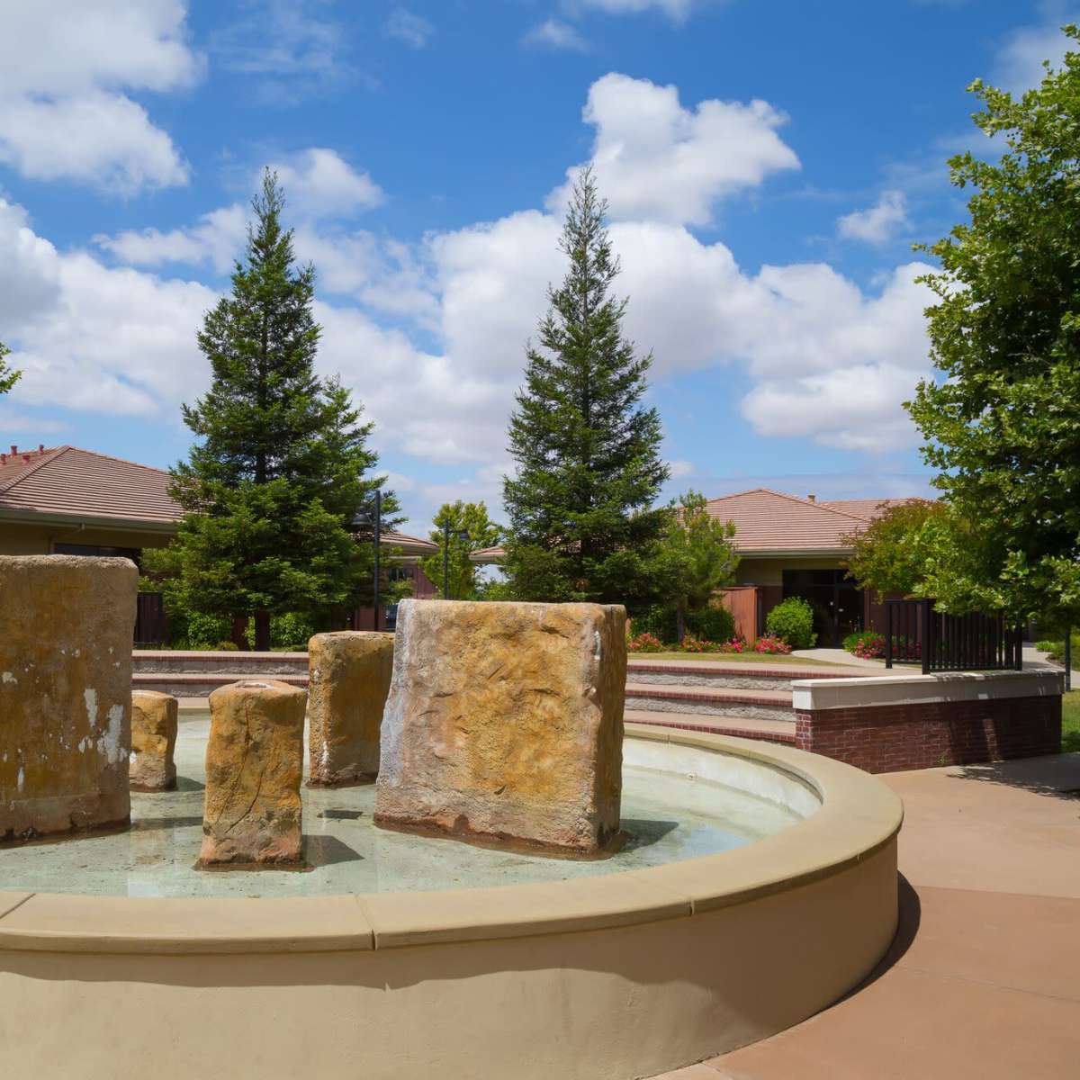 A fountain featuring large stone blocks sits in a landscaped area with green trees and residential buildings in the background.