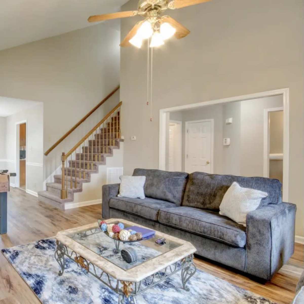 A living room with a gray sofa, a coffee table with decorative items, and a staircase leading to another level, adjacent to a dining area.