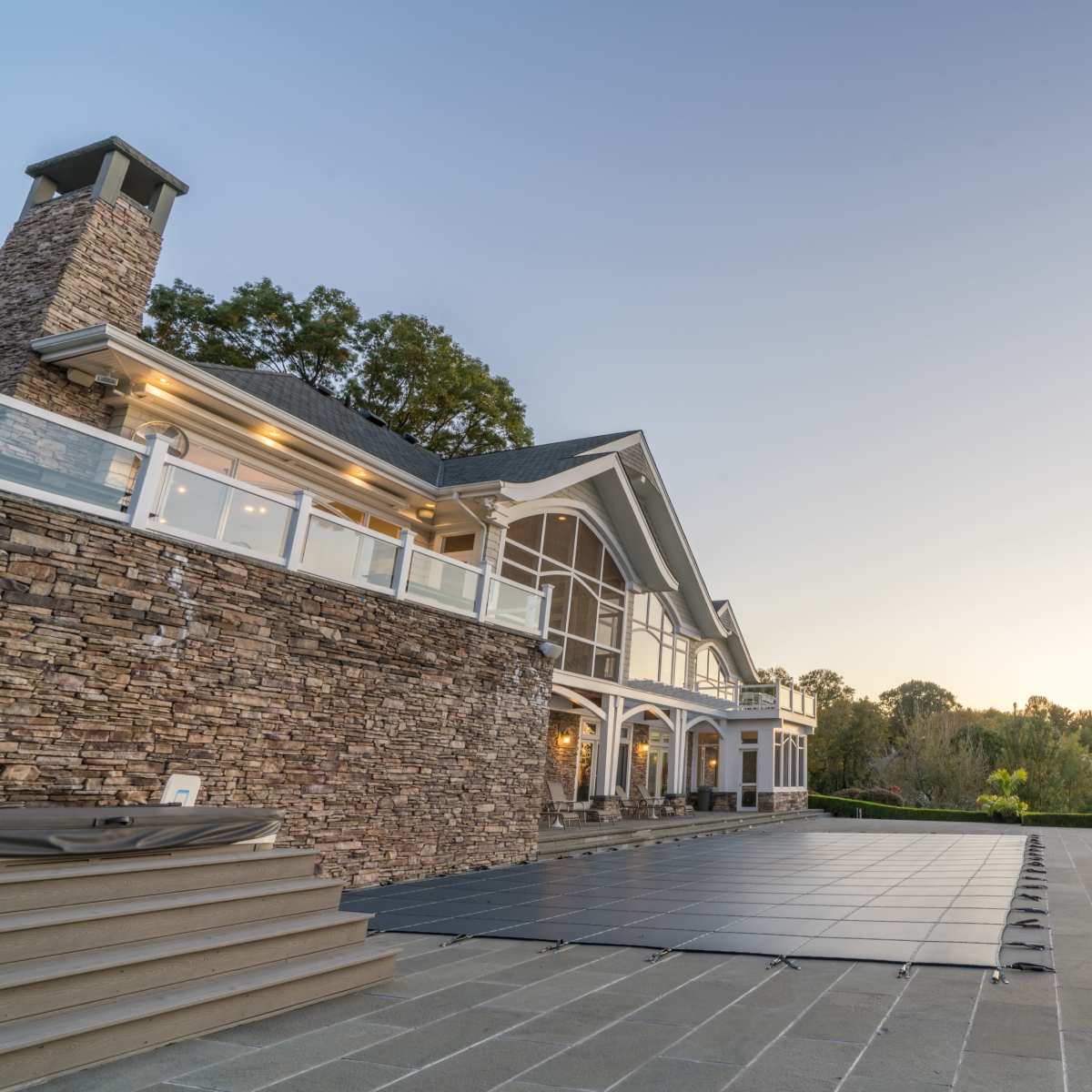 A large stone house with multiple windows overlooks a covered swimming pool and several lounge chairs on a patio.