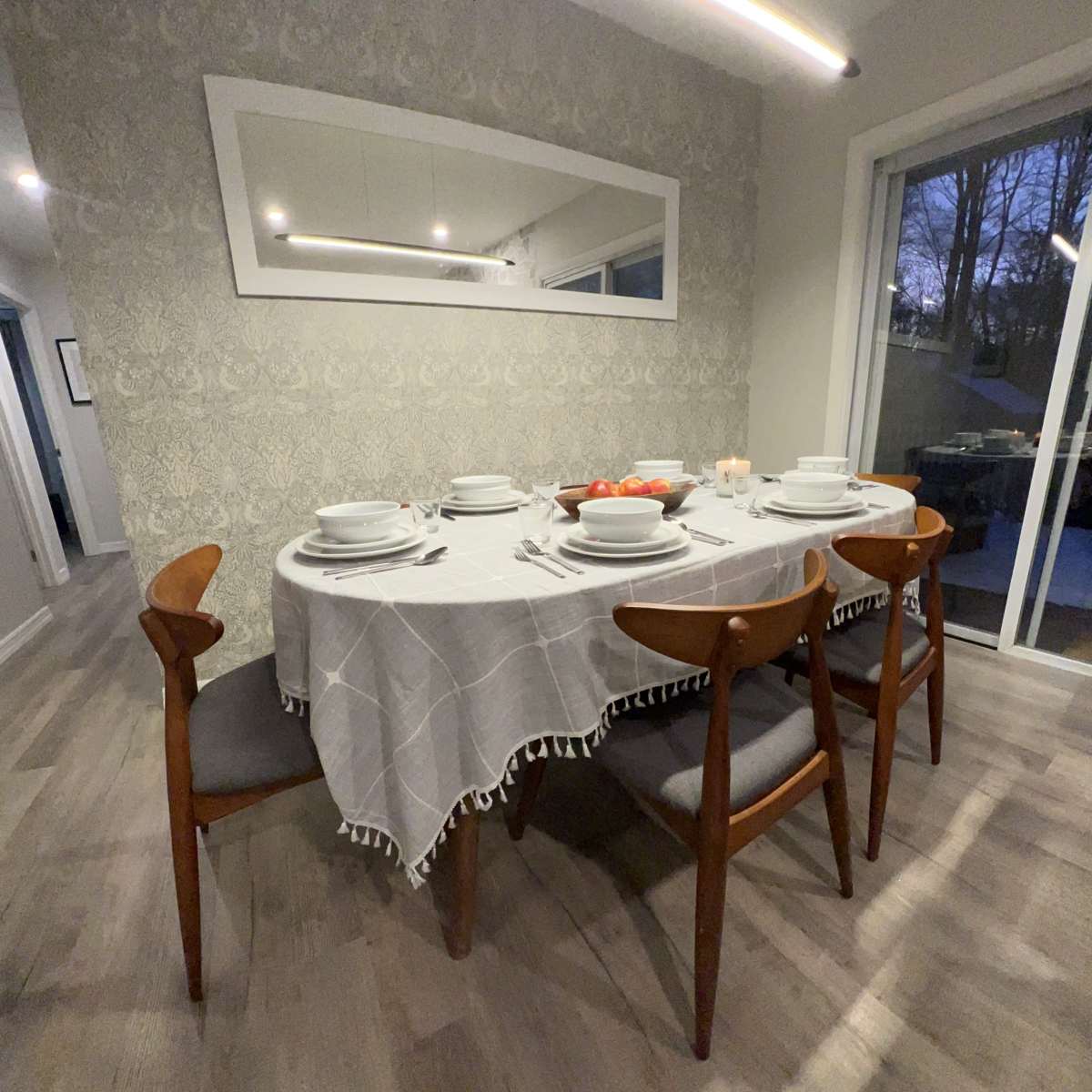 A dining table set with white plates and a light gray tablecloth is arranged with wooden chairs in a well-lit room.