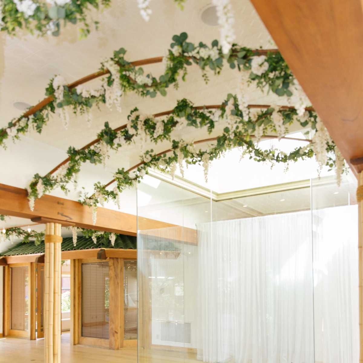 An interior space featuring wooden columns, a glass-walled area with a square cutout in the floor, and greenery along the ceiling.