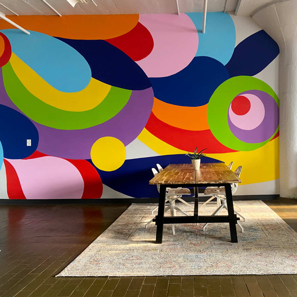 A wooden dining table with white chairs is set in front of a vibrant, abstract mural on a wall in a contemporary room with polished wooden floors.