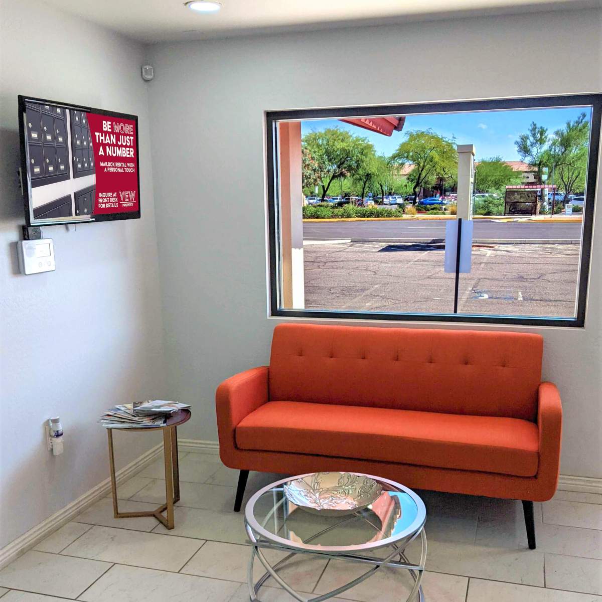 A small waiting area with an orange sofa, a glass coffee table, and a TV mounted on the wall, overlooking a window with a view of the outdoors.