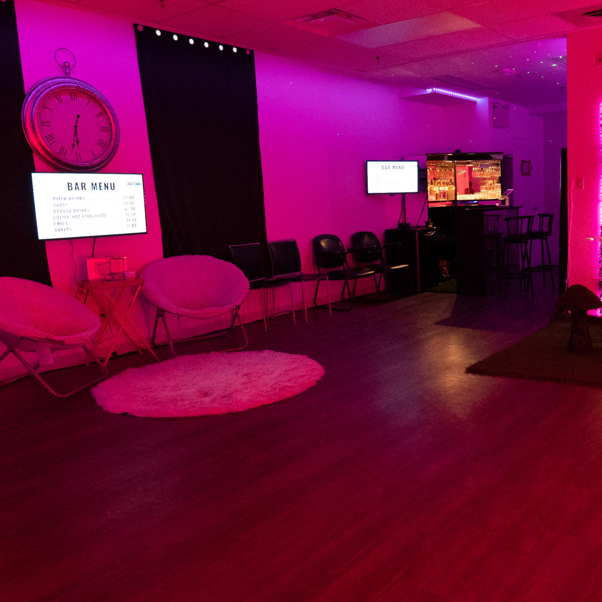 A modern interior space features pink lighting, a bar area with a menu display, and a seating arrangement with chairs and decorative elements.