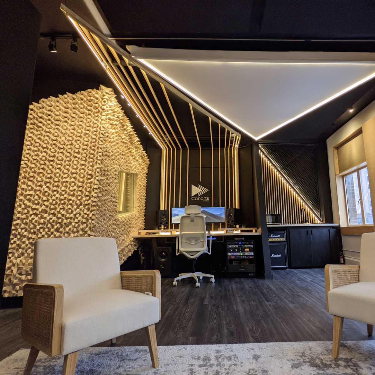 A modern office space with two white chairs facing a sleek desk setup featuring a computer and musical equipment, against a backdrop of geometric wall designs.