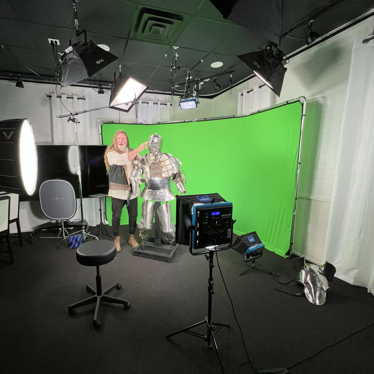 A person adjusts a suit of armor in a studio with a green backdrop and various lighting equipment.