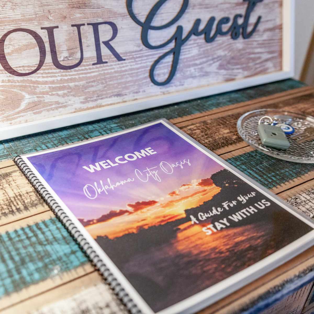 A welcome booklet titled "Oklahoma City Oasis" is placed on a wooden table, alongside a decorative key holder and a sign that says "Be Our Guest."