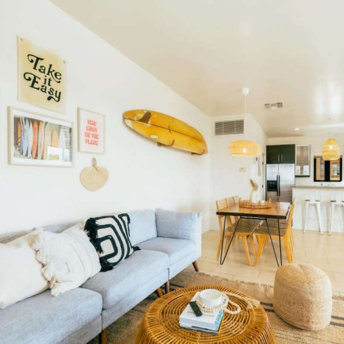 A bright, open-concept living area with a blue couch, a wooden dining table, and a kitchen in the background featuring modern appliances and a surfboard decoration on the wall.