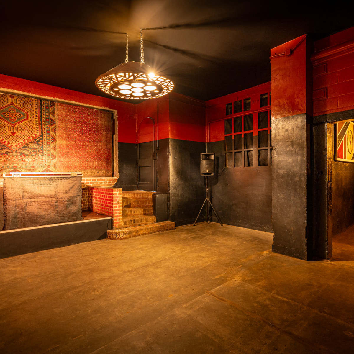 An empty, dimly lit venue featuring a DJ booth with speakers, a patterned rug on the wall, and a staircase leading to a raised area.