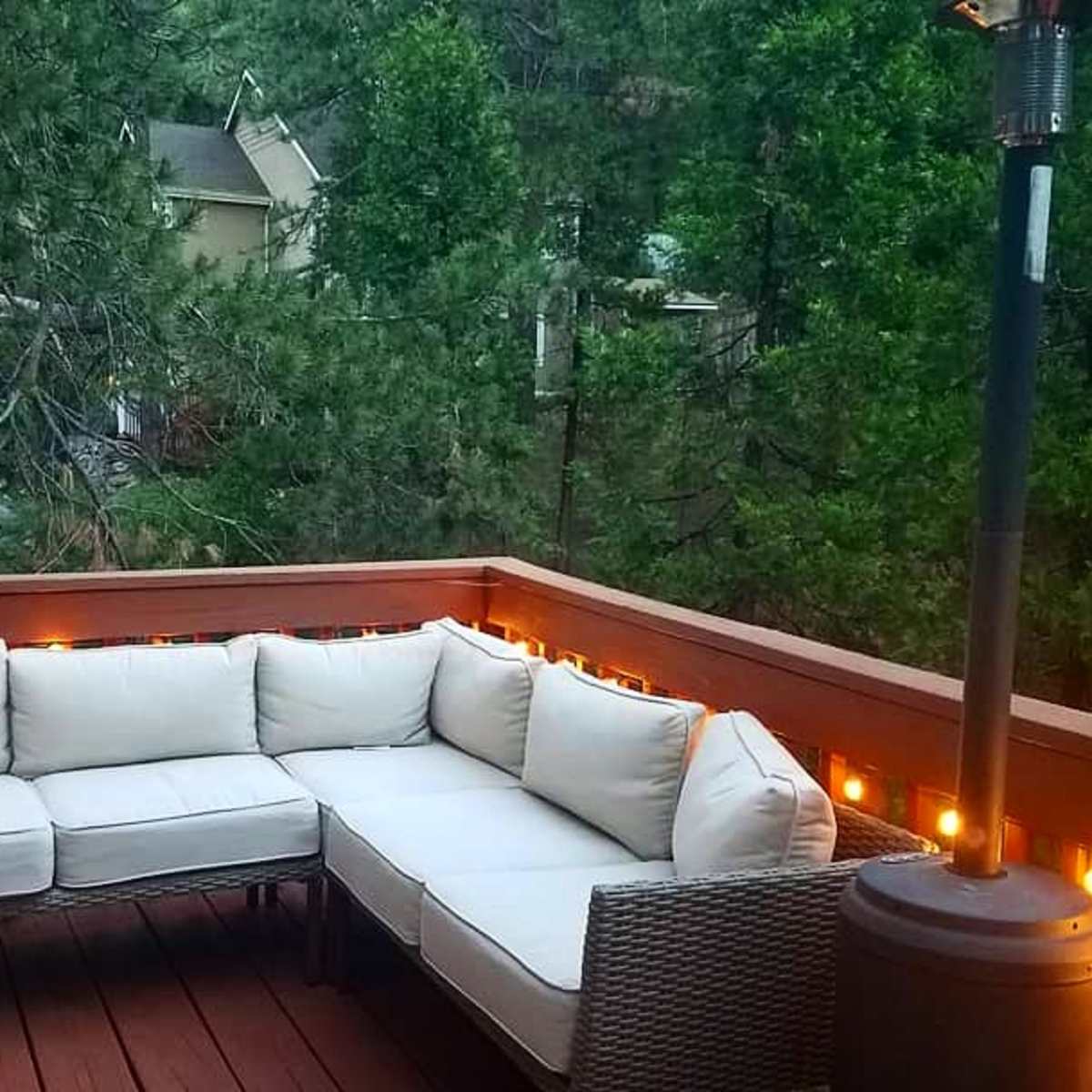 A corner sectional couch with white cushions sits on a wooden deck surrounded by tall trees and string lights.