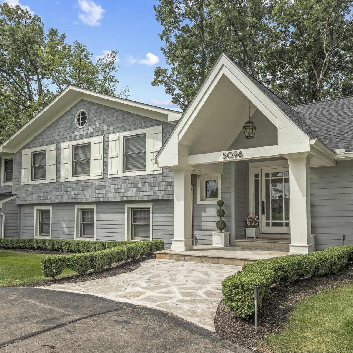 A gray, two-story house with a stone walkway leading to a covered front entrance, surrounded by neatly trimmed bushes and trees.