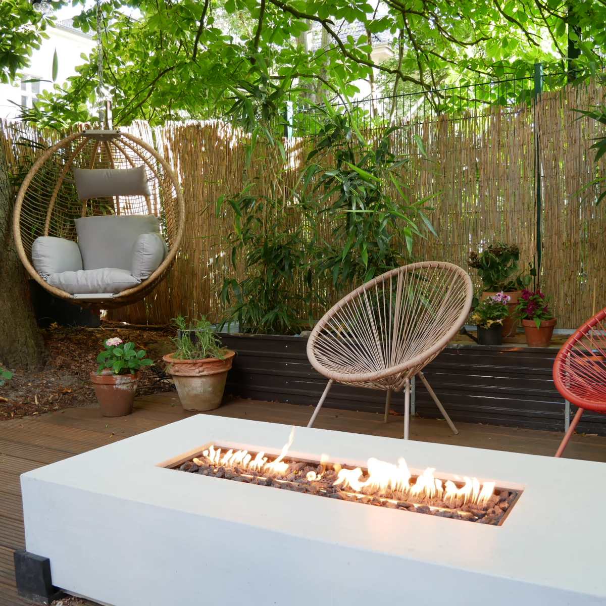 A modern outdoor seating area features a fire pit surrounded by two stylish chairs and a hanging swing, all amidst lush greenery.