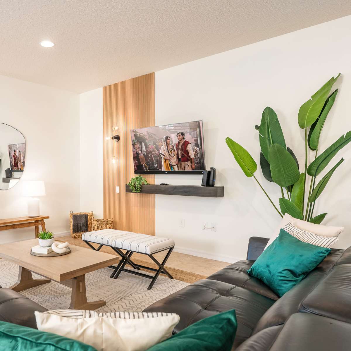 The living room features two dark leather sofas, a wooden coffee table, a large wall-mounted television, and decorative plants, all arranged around a light-colored area rug.