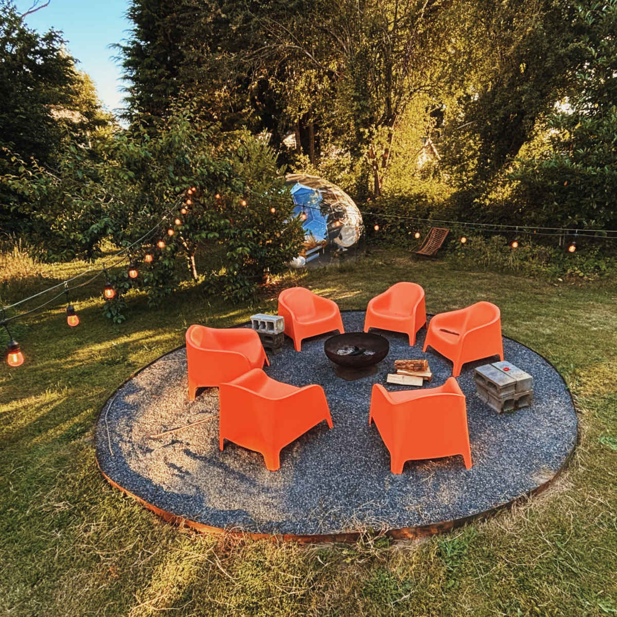 A circular arrangement of five orange chairs surrounds a fire pit on a gravel patio in a grassy yard illuminated by string lights.