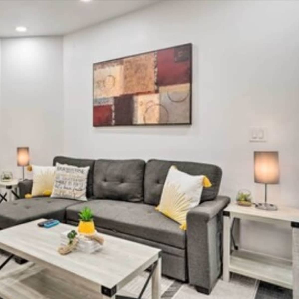 A modern living room features a gray sectional sofa, a coffee table, and decorative lamps, with abstract art on the wall and plants in the corner.