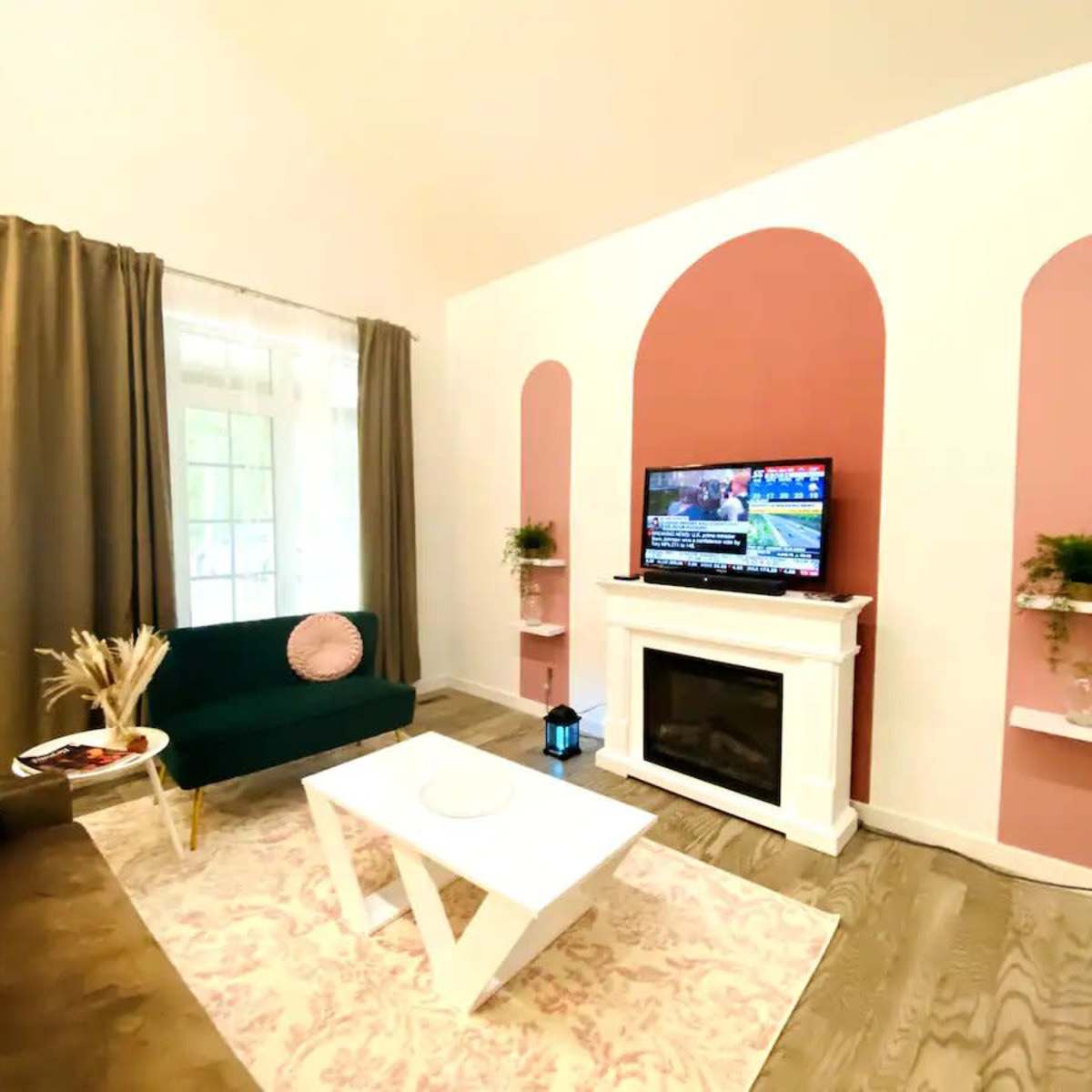 A living room with a green couch, a white coffee table, a television mounted on a pink-accented wall, and decorative plants on shelves.