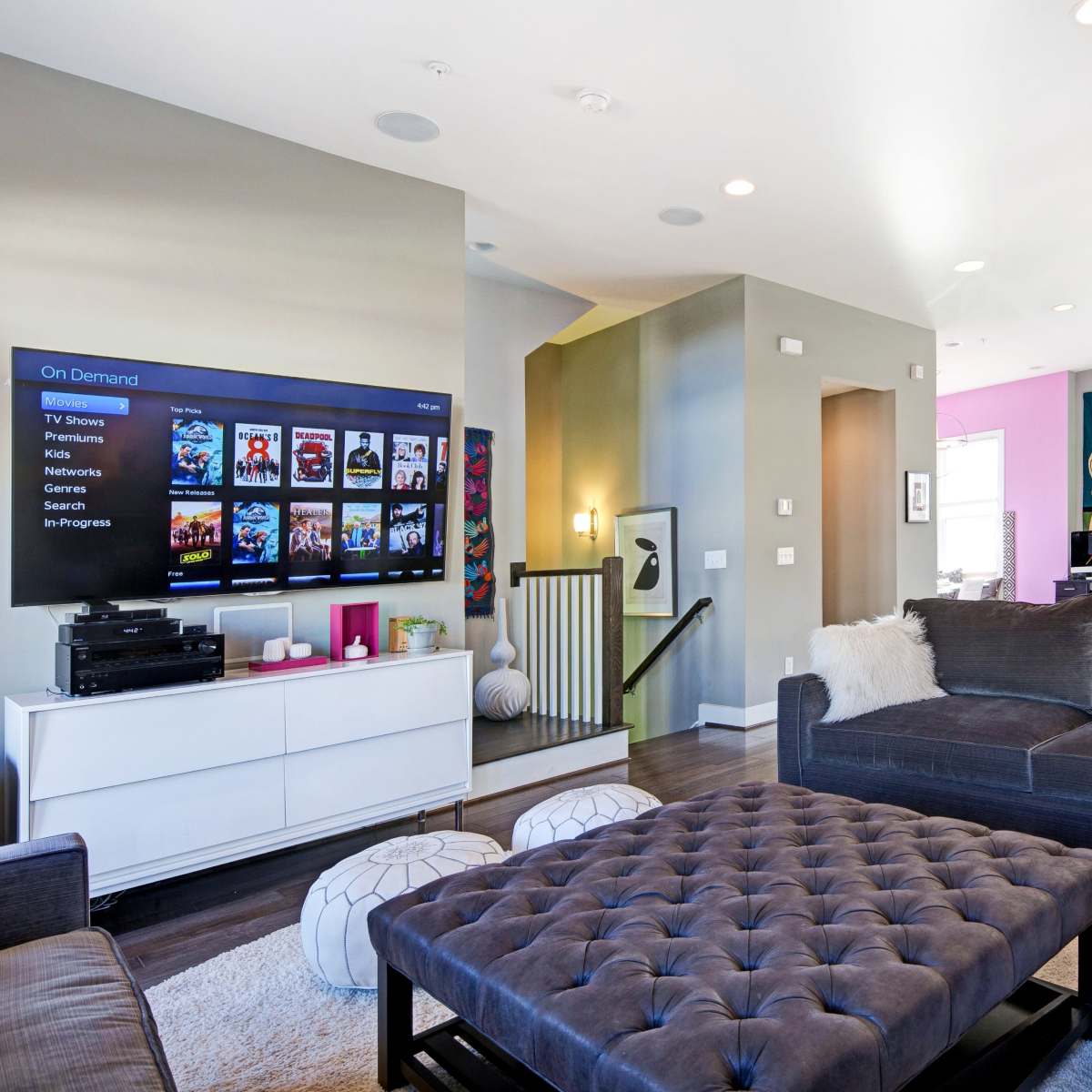 The living room features a large sectional sofa, a tufted coffee table, and a wall-mounted TV displaying on-demand options, with an open view to a kitchen area adorned with colorful decor.