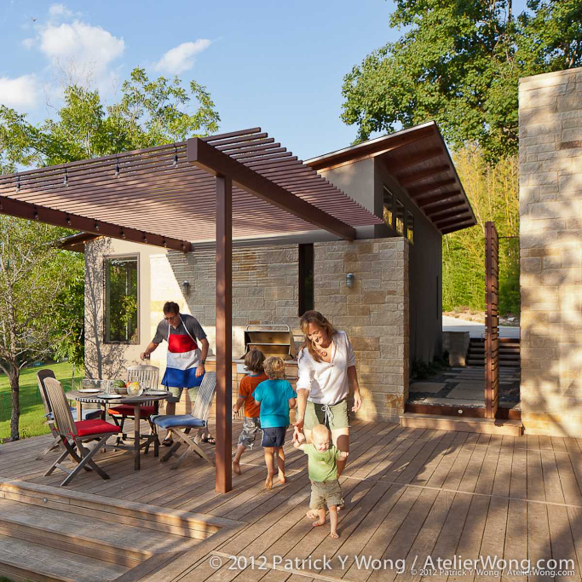 A family gathers on a wooden deck outside a modern home, enjoying a meal under a pergola while children play nearby.
