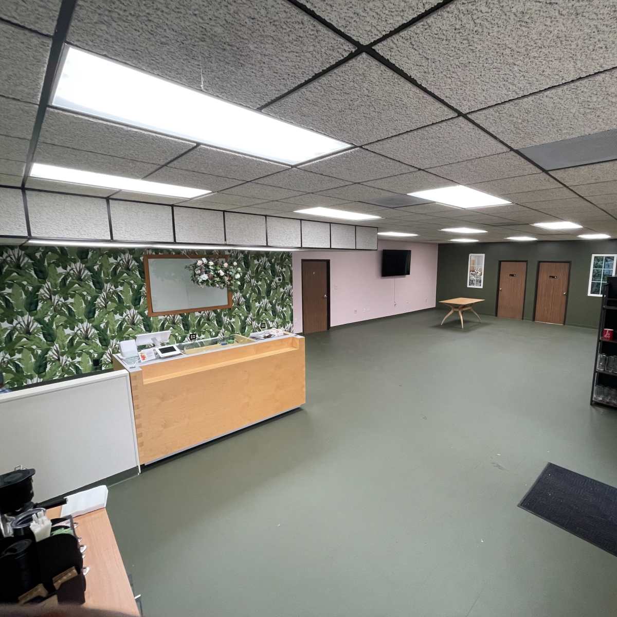 A spacious room with green flooring, a reception area with a counter, a wall decorated with plant-patterned wallpaper, and a central table surrounded by empty space.