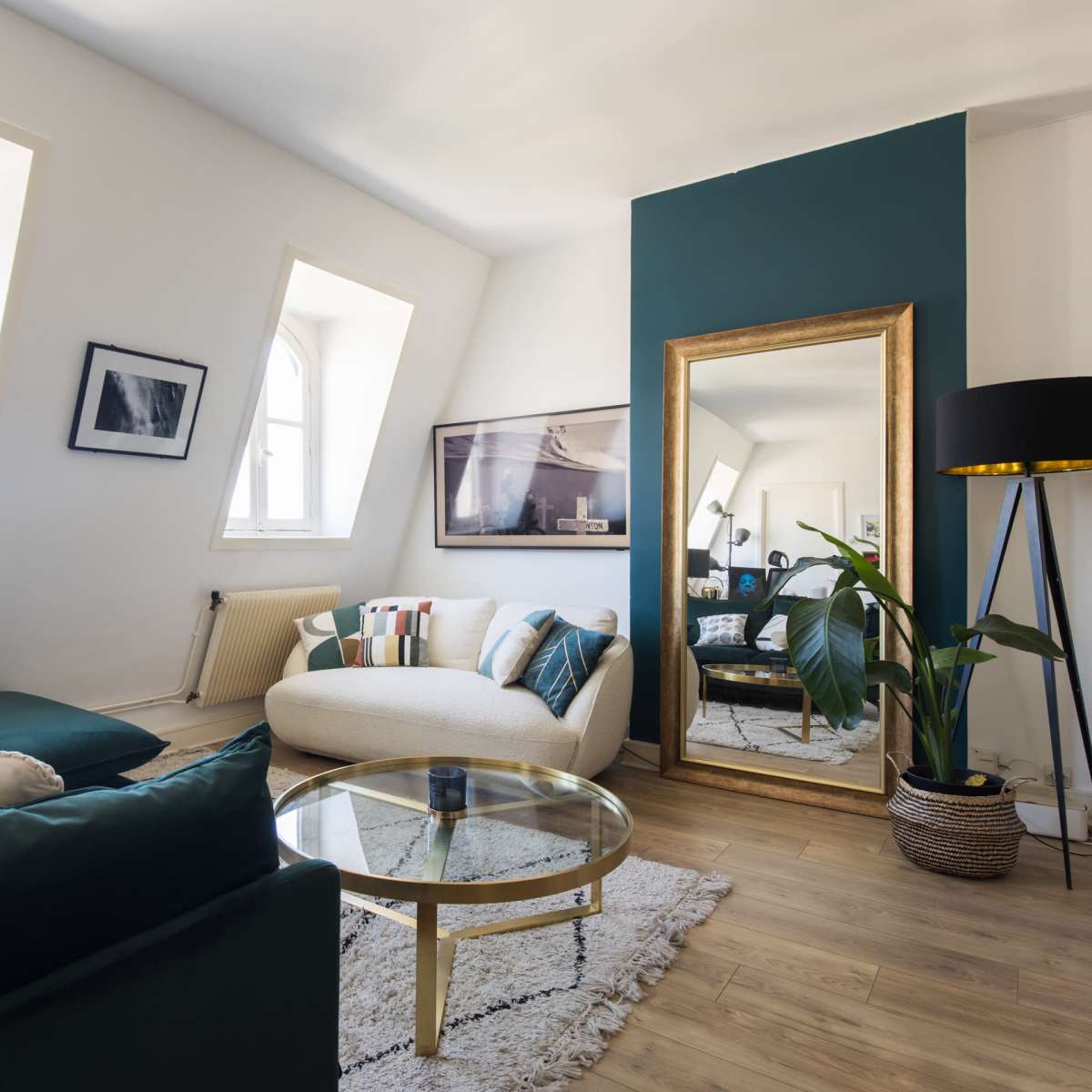 A modern living room features a curved sofa, a coffee table, and a large mirror against a colorful accent wall, with natural light streaming through angled windows.
