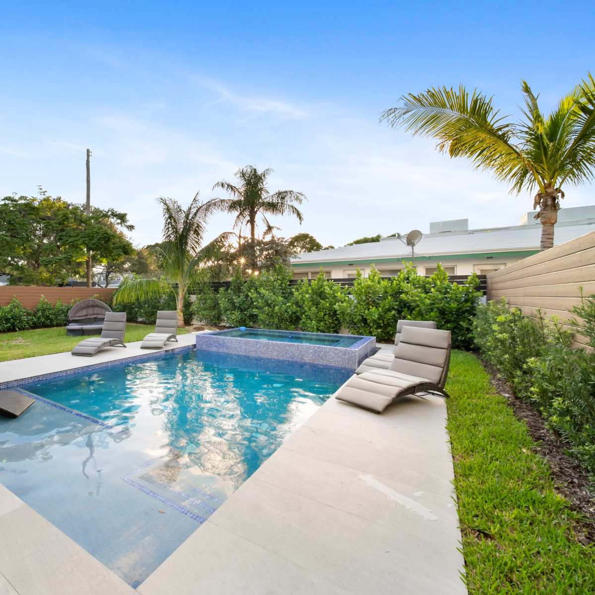 A backyard features a swimming pool with a spa and several lounge chairs surrounded by palm trees and manicured grass.