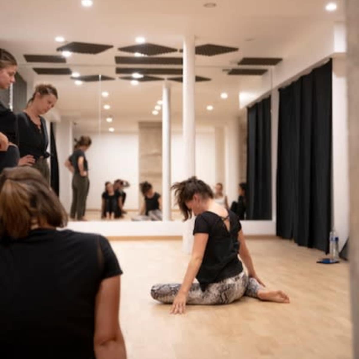 In a dance studio with mirrors, several individuals are observing one person performing a split on the floor.