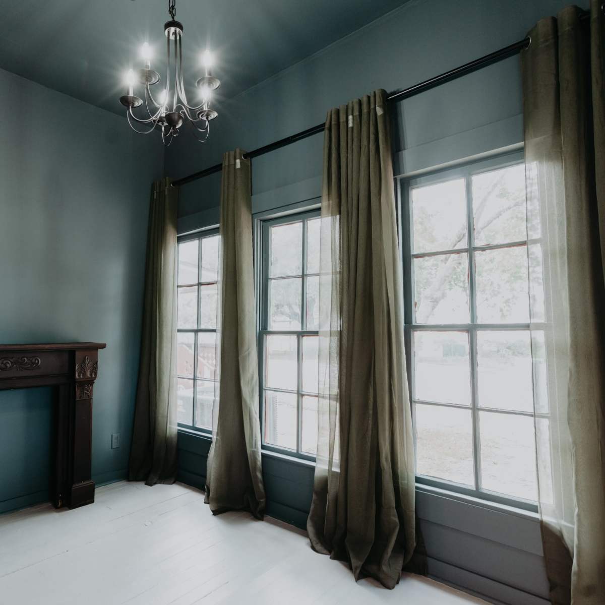 A room with a dark blue wall, a chandelier, and tall windows covered by green curtains, along with a wooden mantel against one wall.