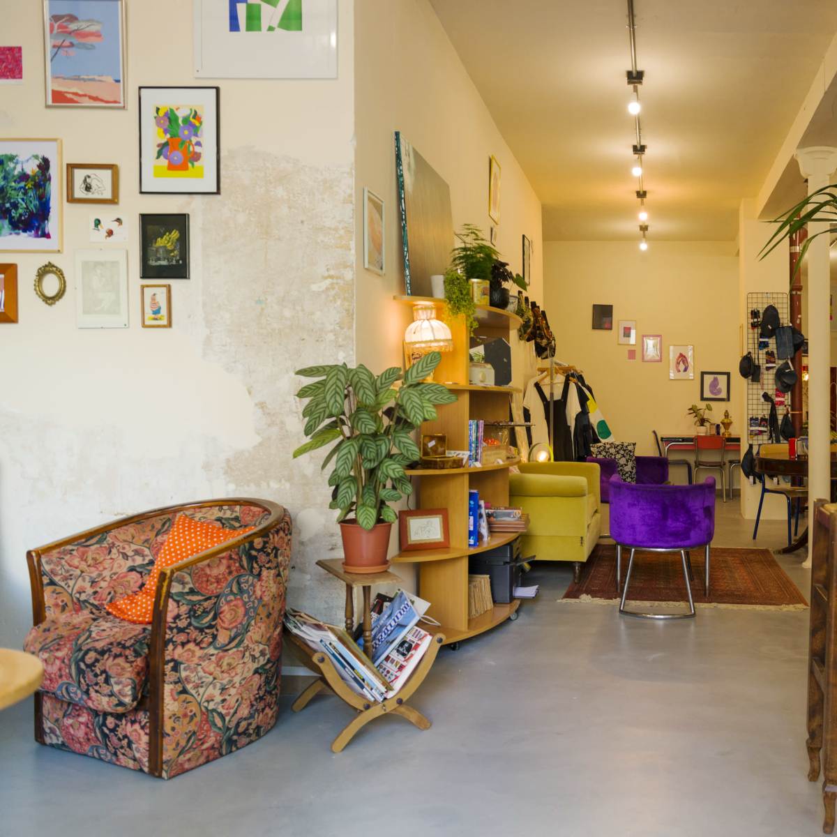 A cozy café with various seating areas, including a patterned armchair and a purple chair, along with plants, artwork on the walls, and a chalkboard menu.