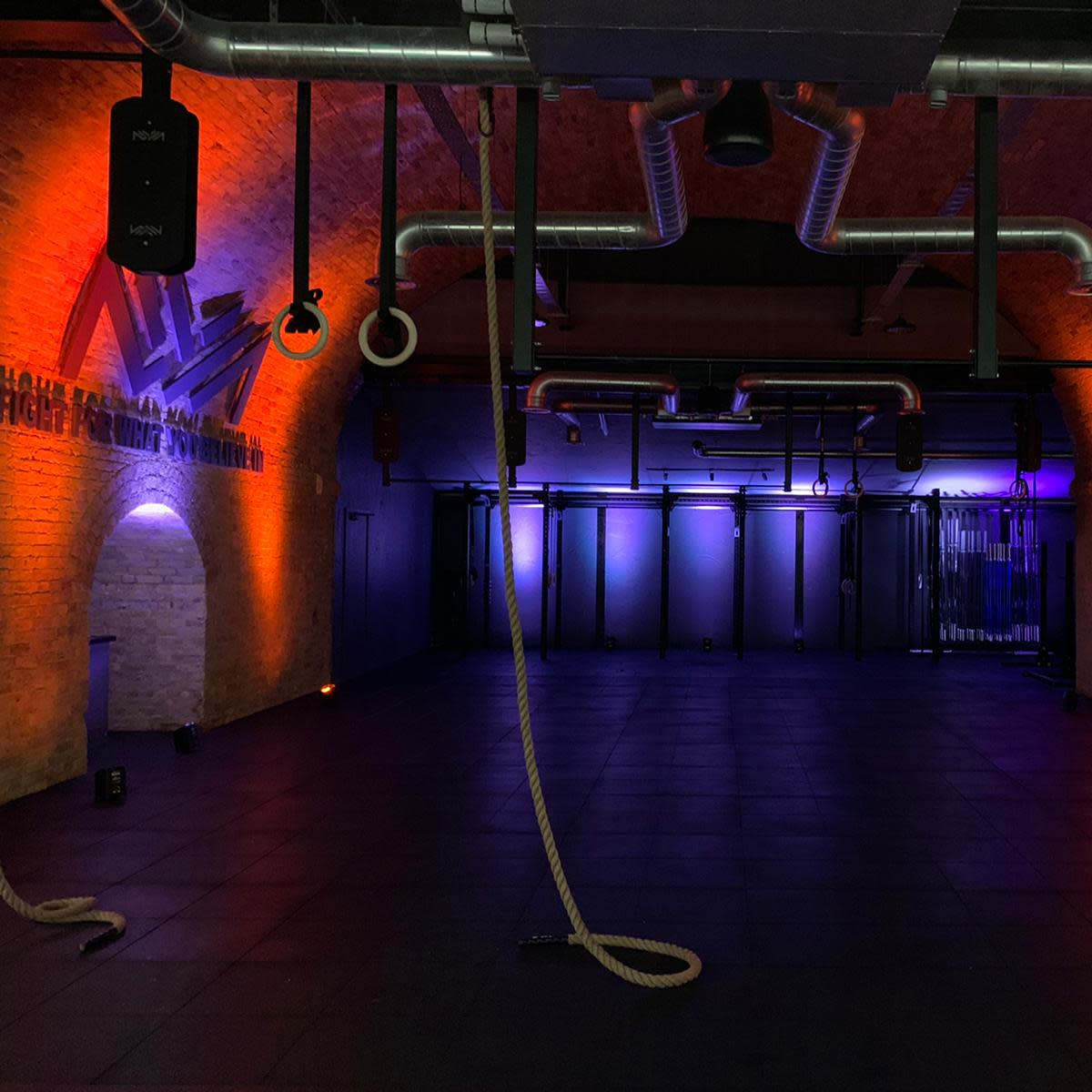 An empty gym interior with exposed brick walls, metallic pipes, and ropes hanging from the ceiling, illuminated by purple and orange lights.