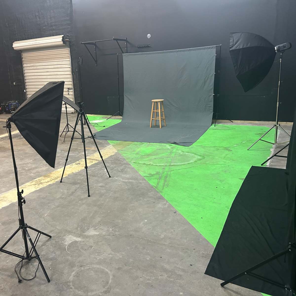 A photo studio setup with a gray backdrop, a wooden stool in the center, and multiple lighting umbrellas on stands, against a dark wall and concrete floor.
