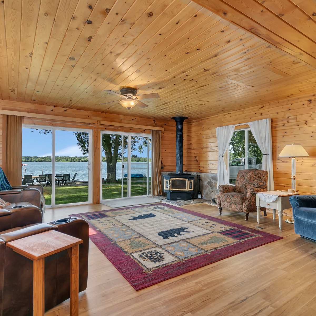 The room features wooden walls and ceiling, a fireplace, and large windows overlooking a lake.