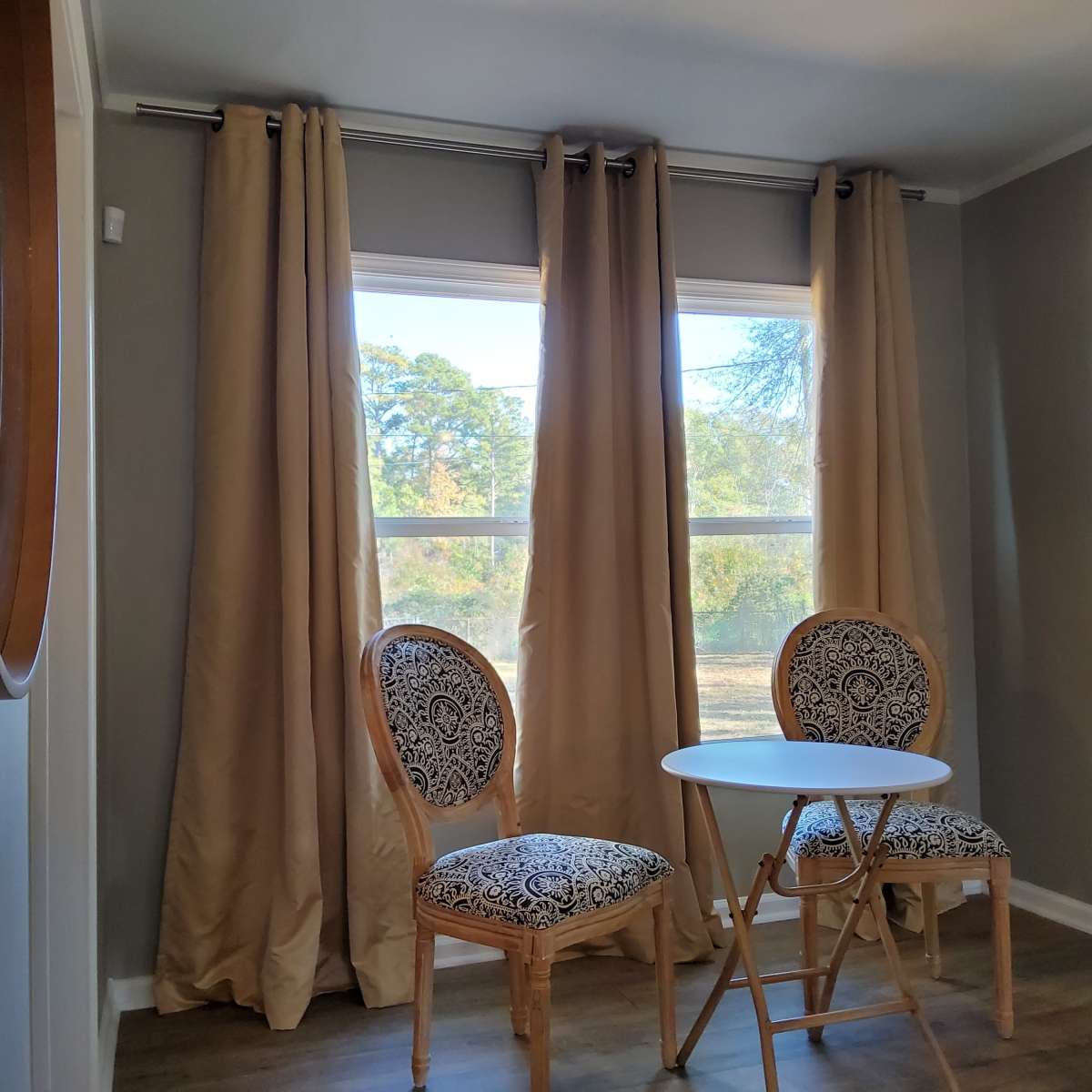 A small seating area with two patterned chairs positioned around a white circular table, set against a backdrop of large windows with beige curtains.