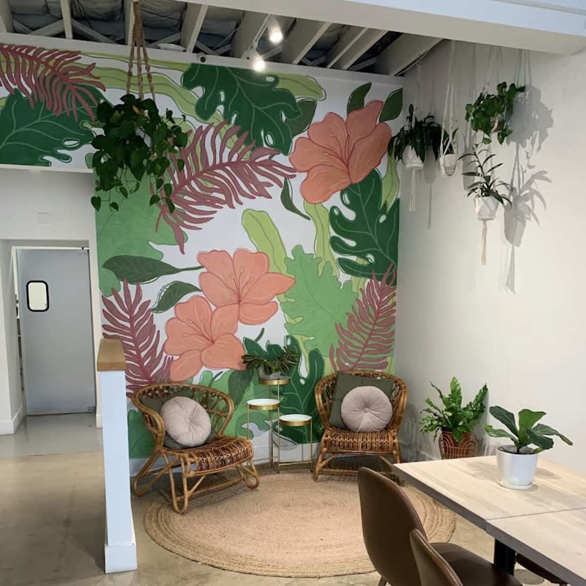 A cozy corner of a cafe featuring a tropical-themed mural, two rattan chairs, a small table, and various plants in pots.