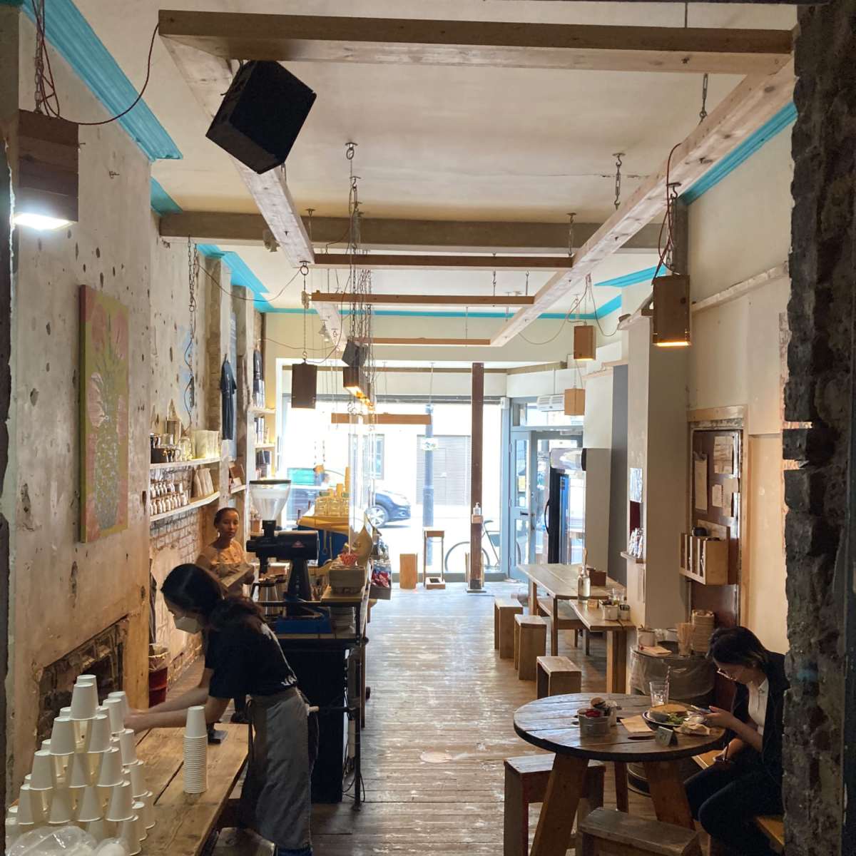 The interior of a coffee shop features wooden tables, a counter with a barista, and a series of stools against stone walls.