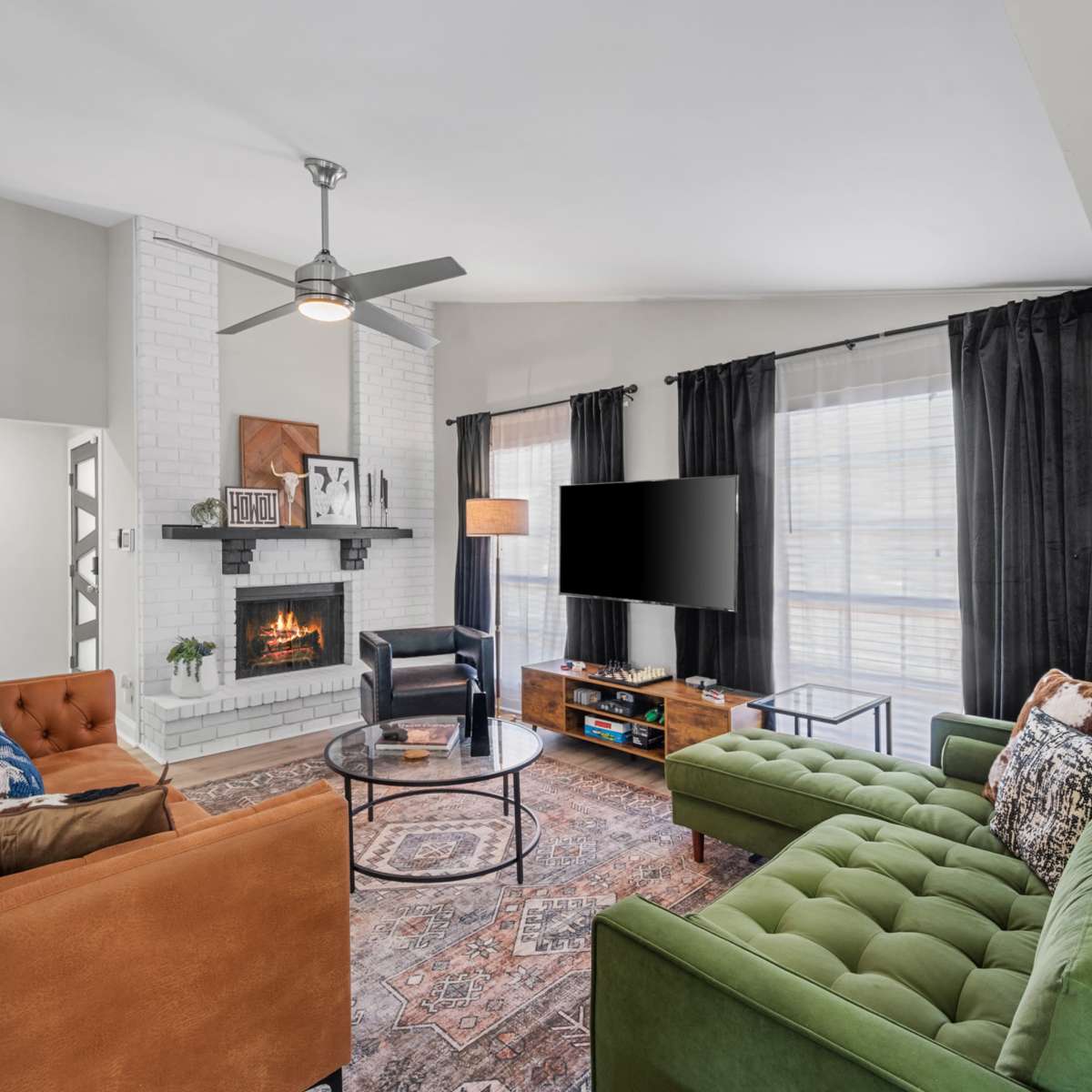 A living room features a cozy seating arrangement with an orange and green couch, a round coffee table, a white brick fireplace, and a television mounted on the wall.