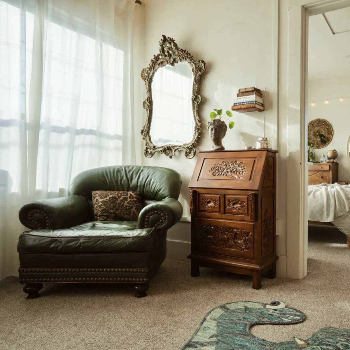 A cozy room with a green armchair, an ornate mirror, a wooden chest, and a bed visible through an open doorway, surrounded by indoor plants.