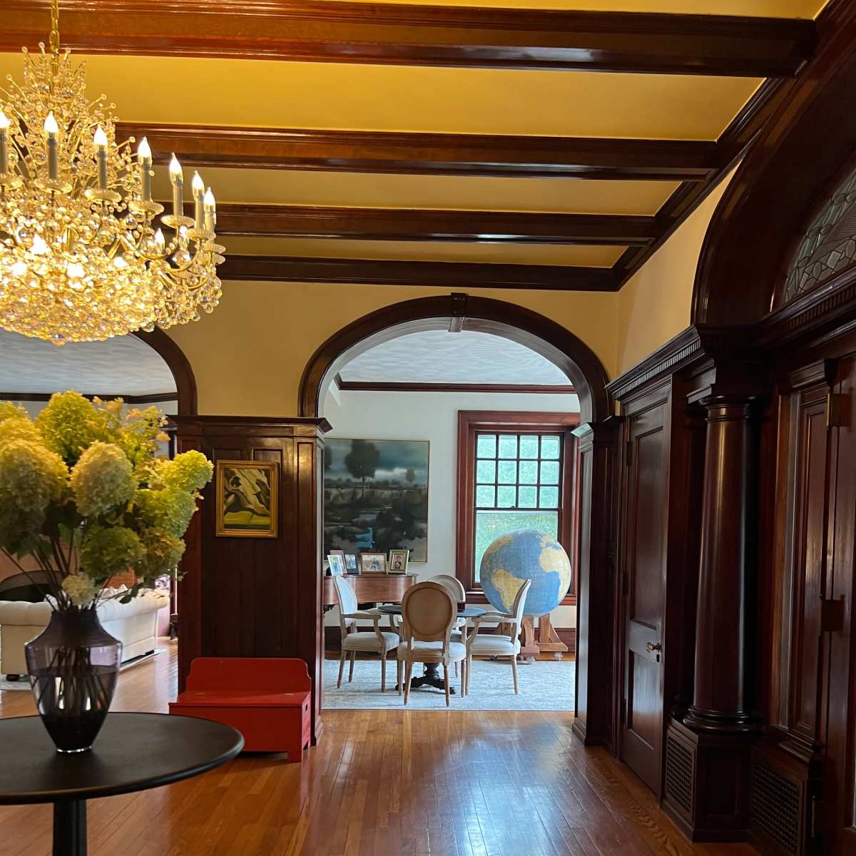 A hallway with a chandelier and a floral arrangement leads to a sitting area featuring a globe and elegant furniture.