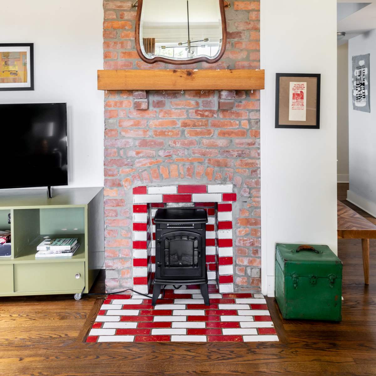 A living room featuring a red and white striped fireplace, a wall-mounted mirror above it, a TV on a green media console, and a vintage green chest.