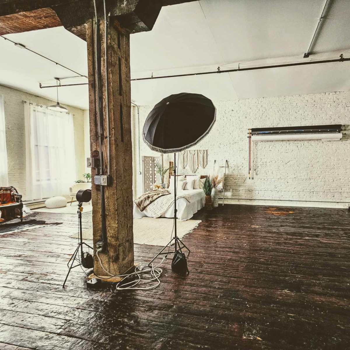 A spacious, well-lit bedroom with wooden floors, a large bed, and a photography setup including a softbox and a backdrop.