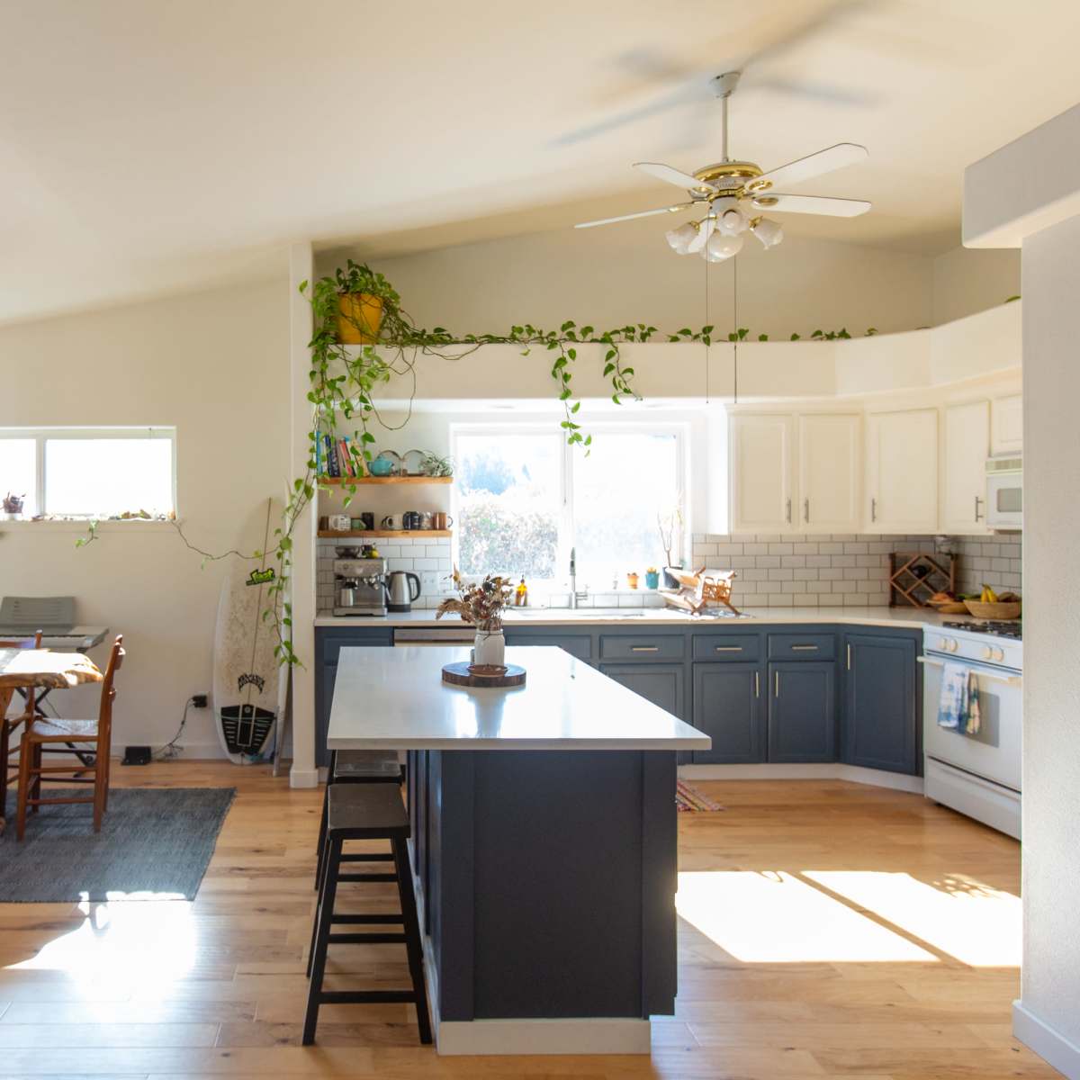 A bright kitchen features a central island, white cabinetry, and a dining area with wooden chairs and a table near a large window.