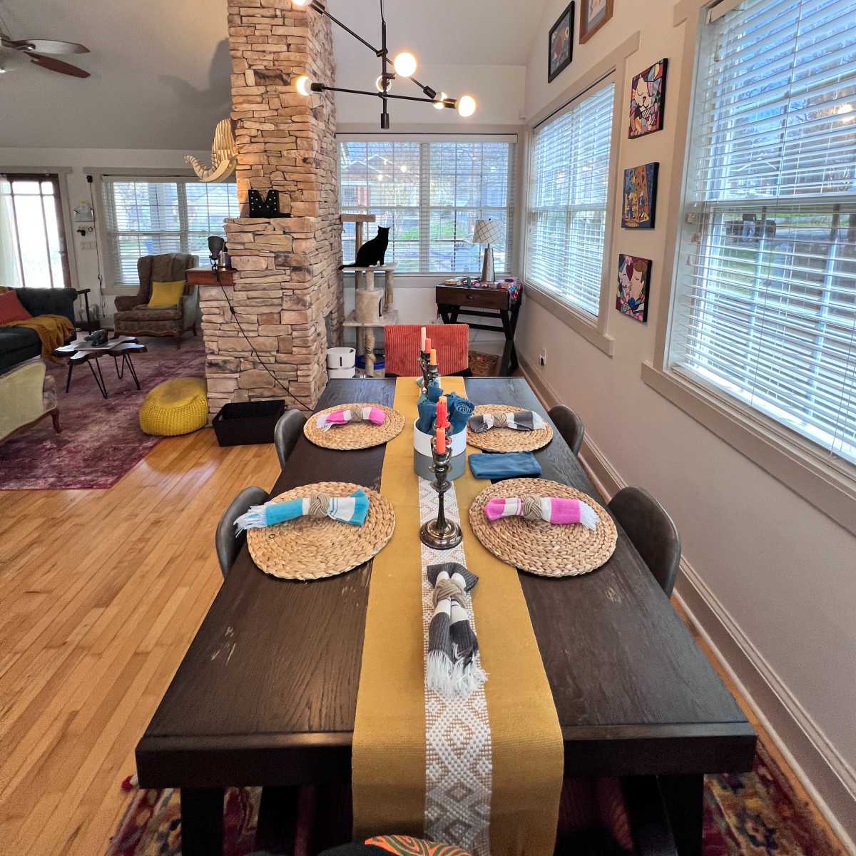 A dining table set with place settings and decorative items is positioned beside a stone column in a brightly lit room featuring large windows and various artworks on the walls.