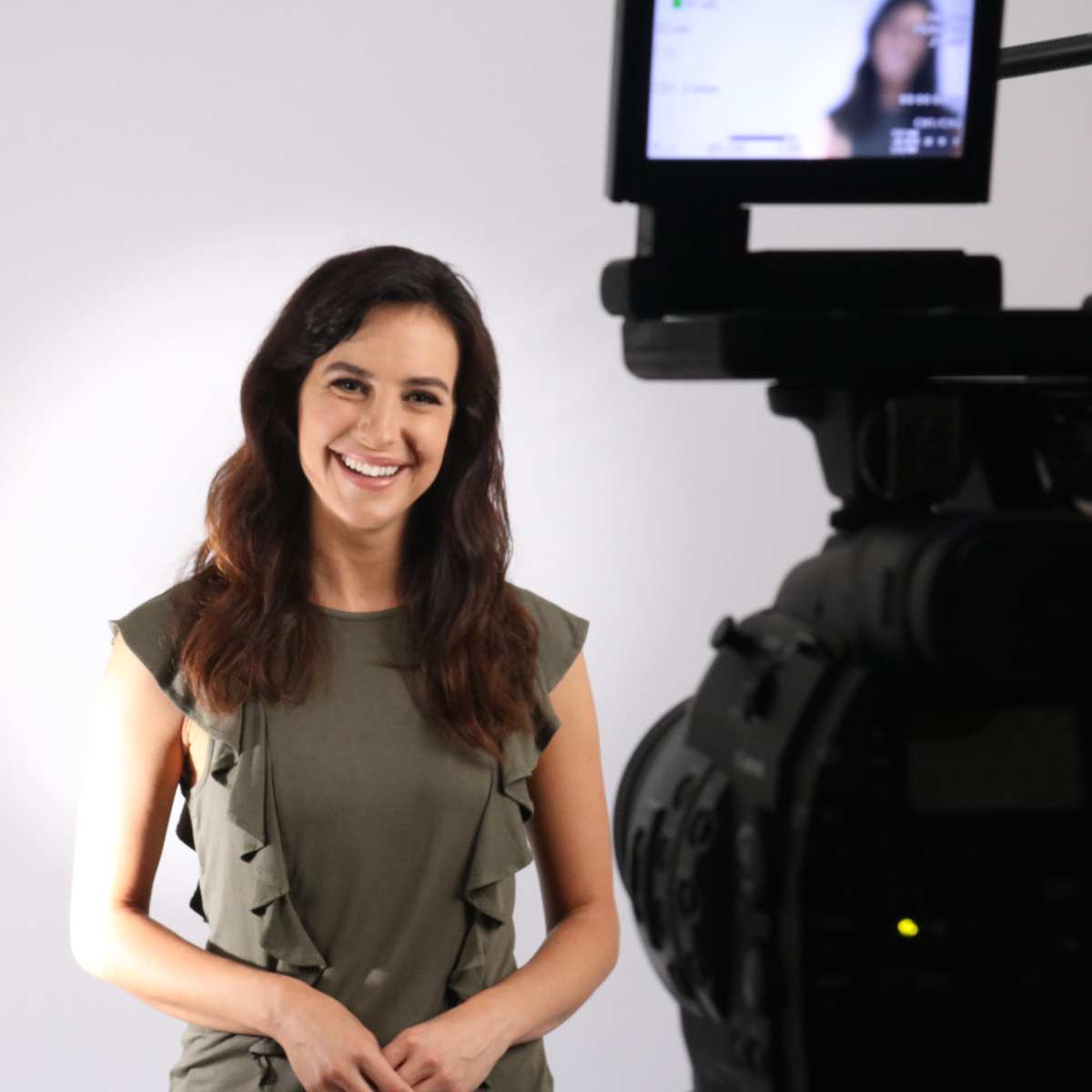 A woman stands in front of a neutral background, smiling while facing a camera on a tripod.