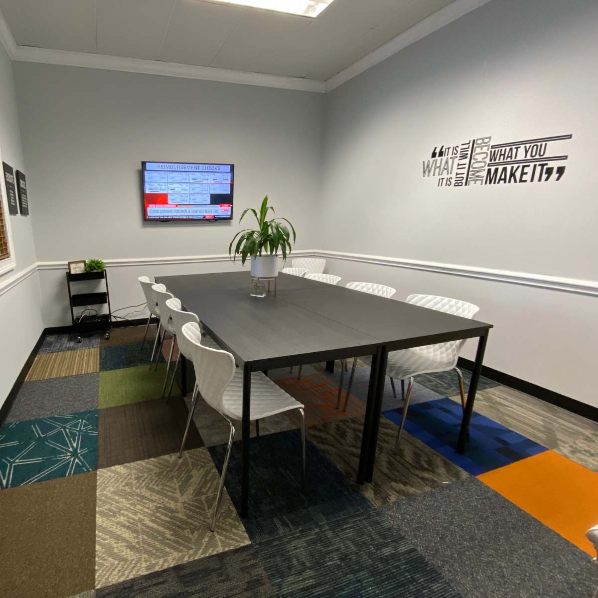 A modern conference room with a rectangular table surrounded by white chairs, a television on the wall, and a decorative plant.
