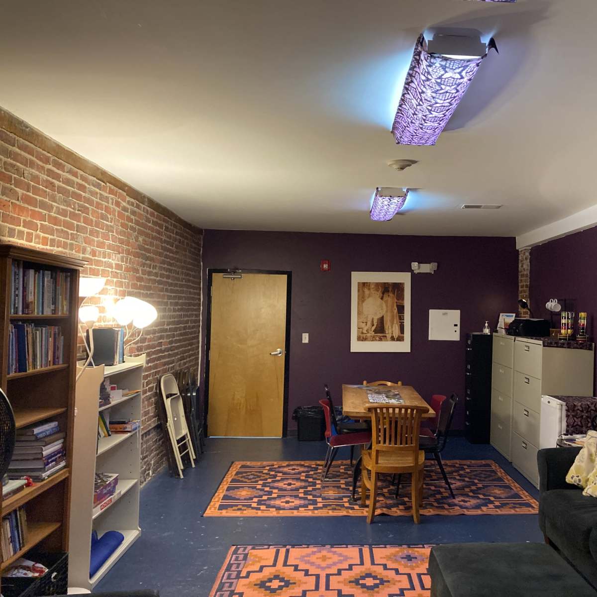 A room with purple walls, a brick wall section, a table with chairs, a couch, and shelving filled with books.