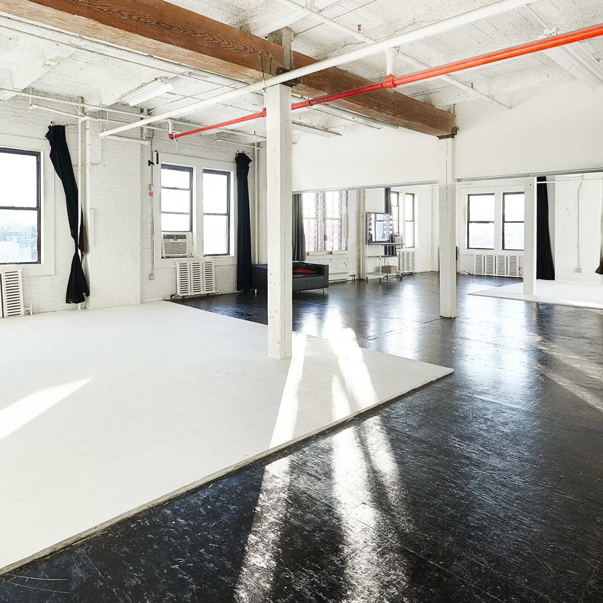 A spacious, industrial-style room with white walls, large windows, and a black polished floor, featuring a seamless backdrop area on one side.